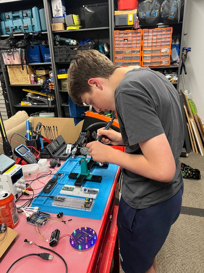 My son soldering the BSides Canberra 2026 badge at the electronics workbench in the shed