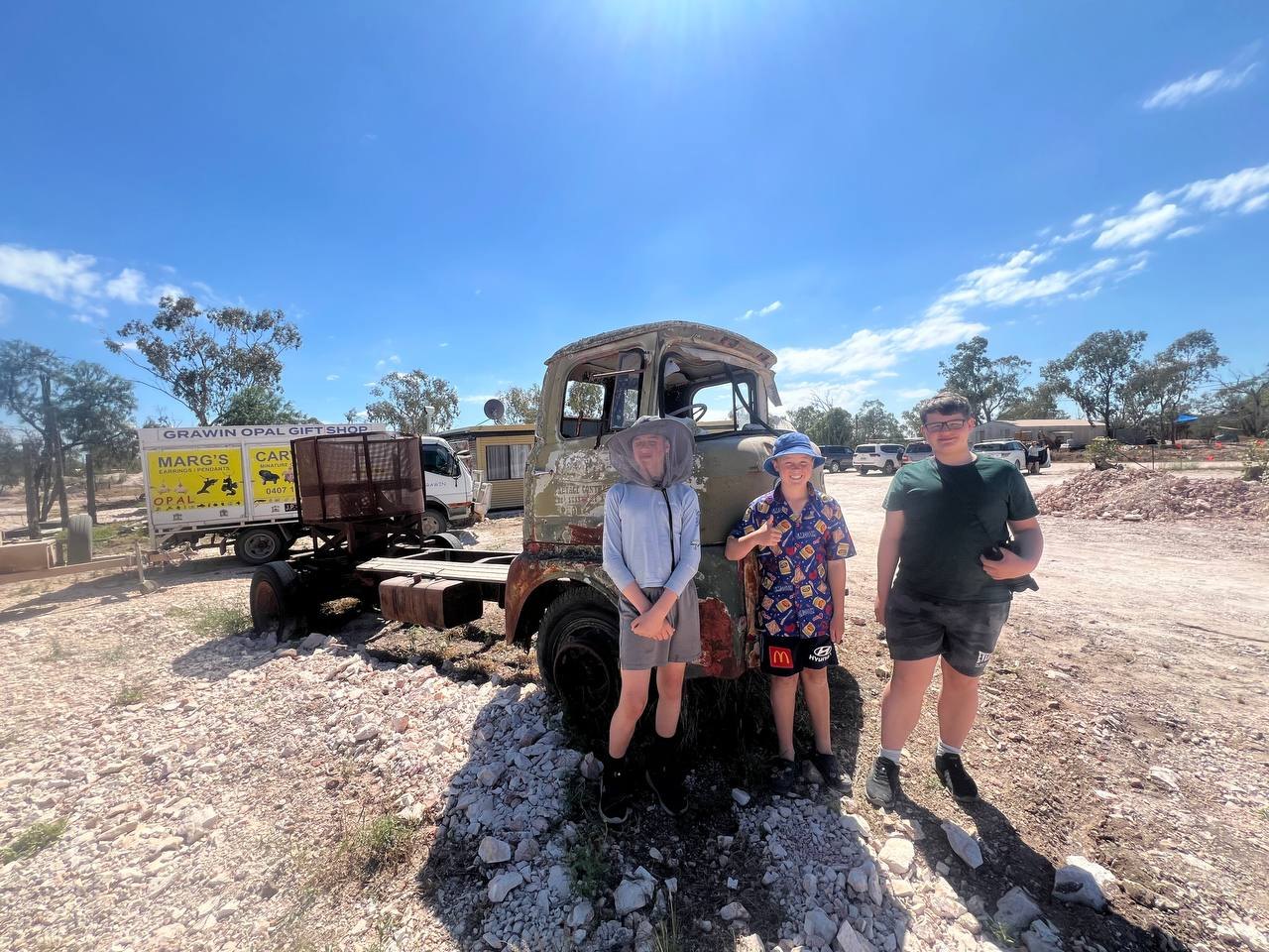 Three of the kids in front of a rusty truck, under signs for the Grawin Opal Gift Shop and Marg's Opal