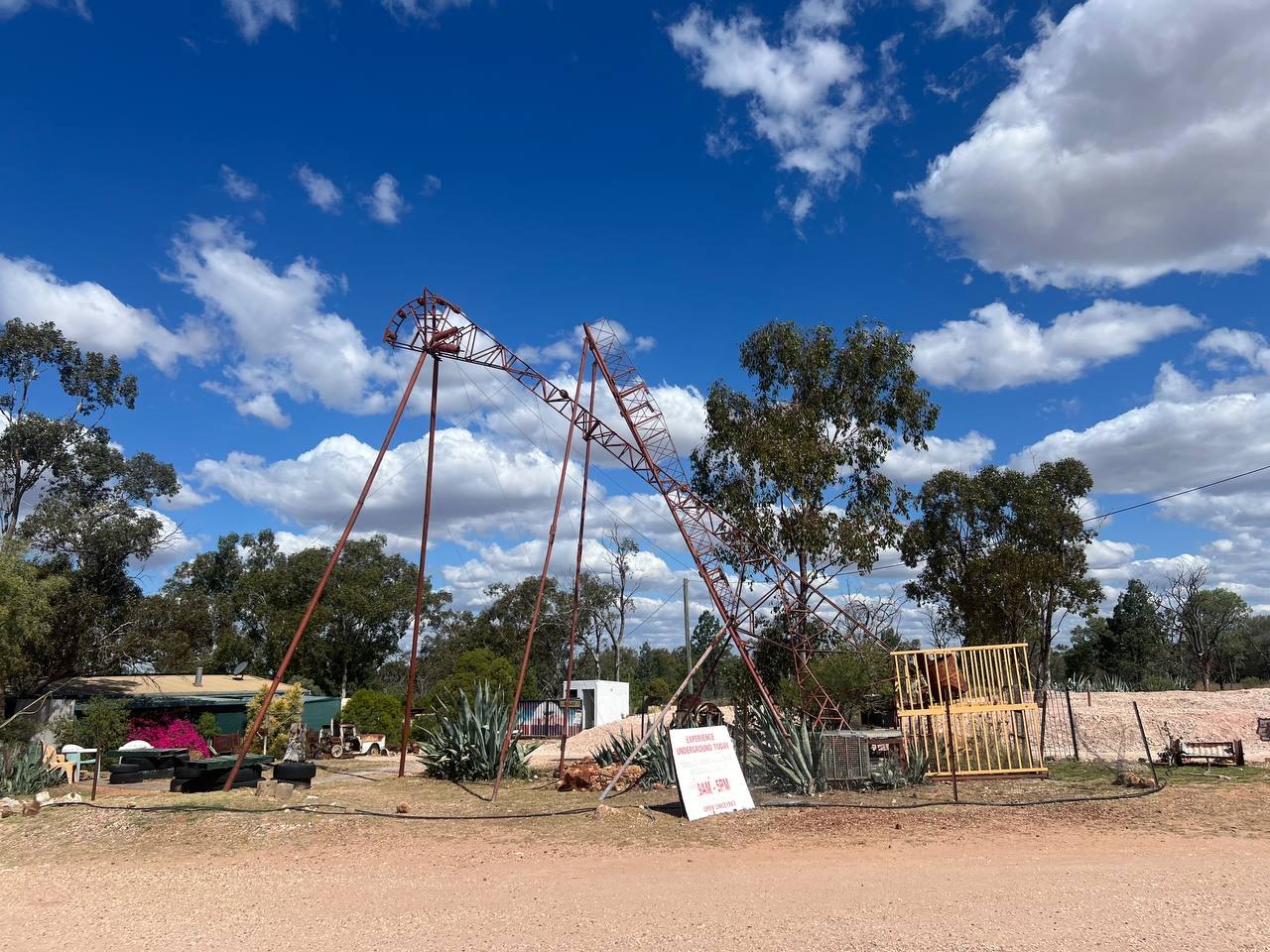 A rusted steel mine headframe and hoist in a gravel yard, with a sign advertising underground tours from 9am to 5pm