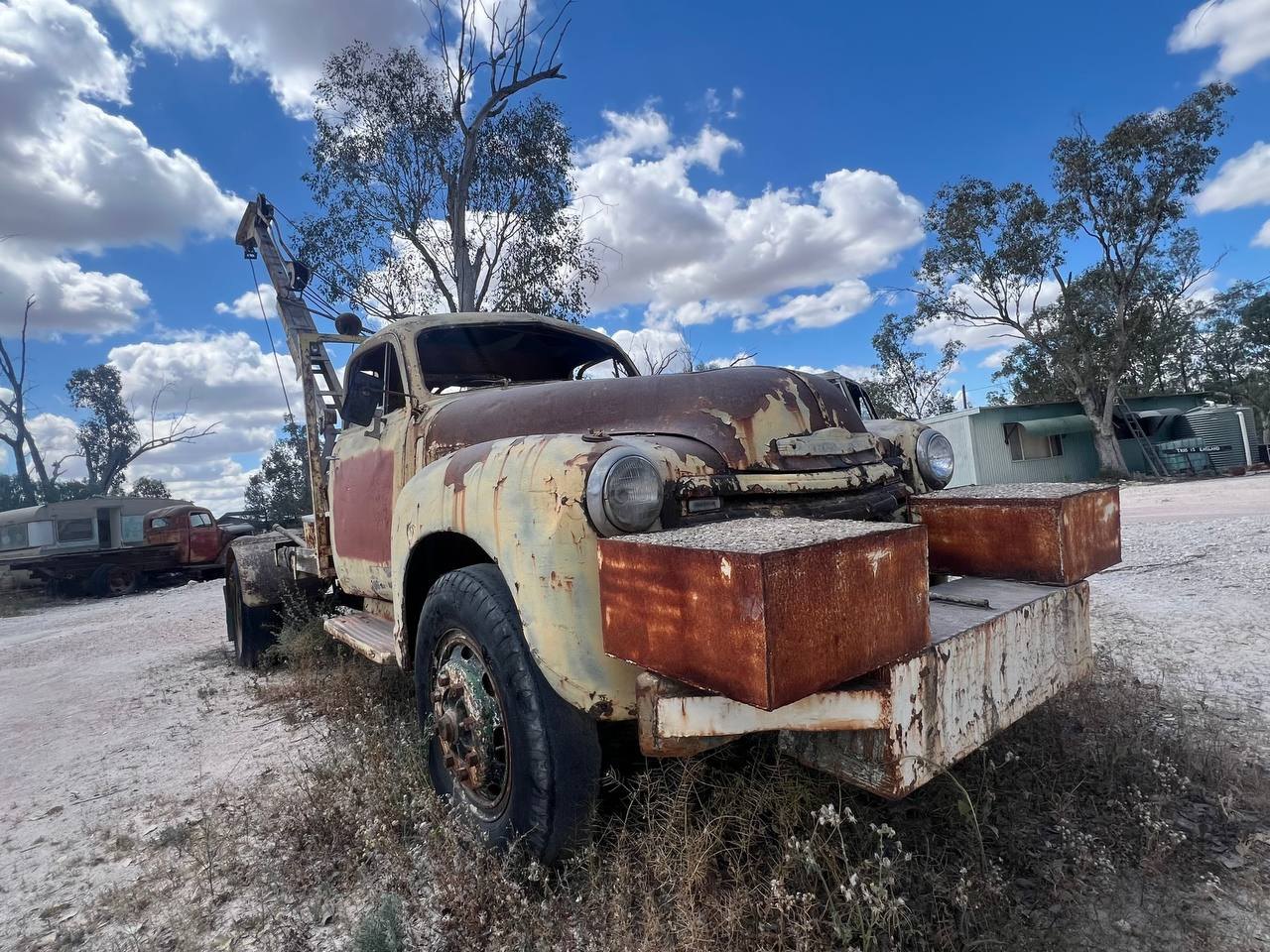 A heavily rusted vintage tow truck with a crane rig, cream and maroon paint peeling, in a yard of derelict vehicles