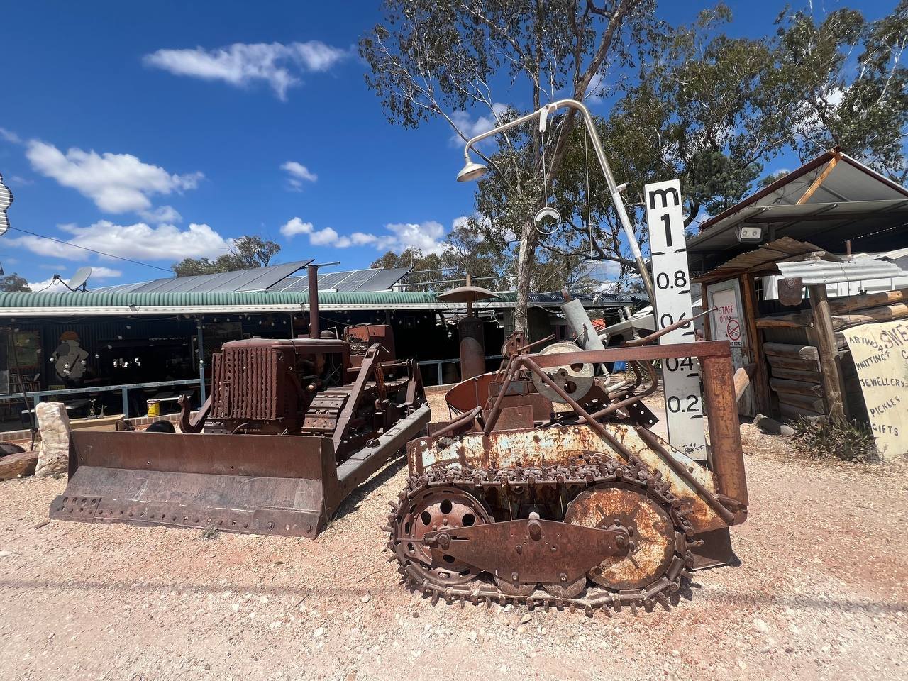 Rusted vintage bulldozer and tracked mining vehicle on gravel in front of sheds, with a sign listing artwork, jewellery, pickles, and gifts